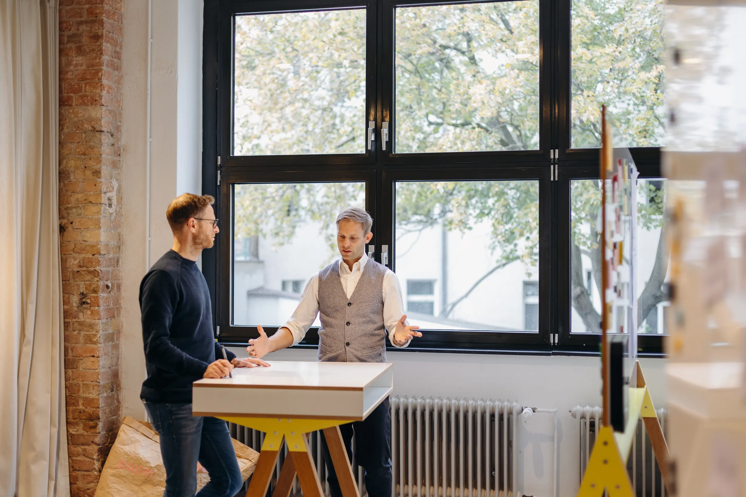Kevin Rassner (mit Weste) im Coaching-Gespräch mit einer Führungskraft an einem Stehtisch vor einem großen Fenster