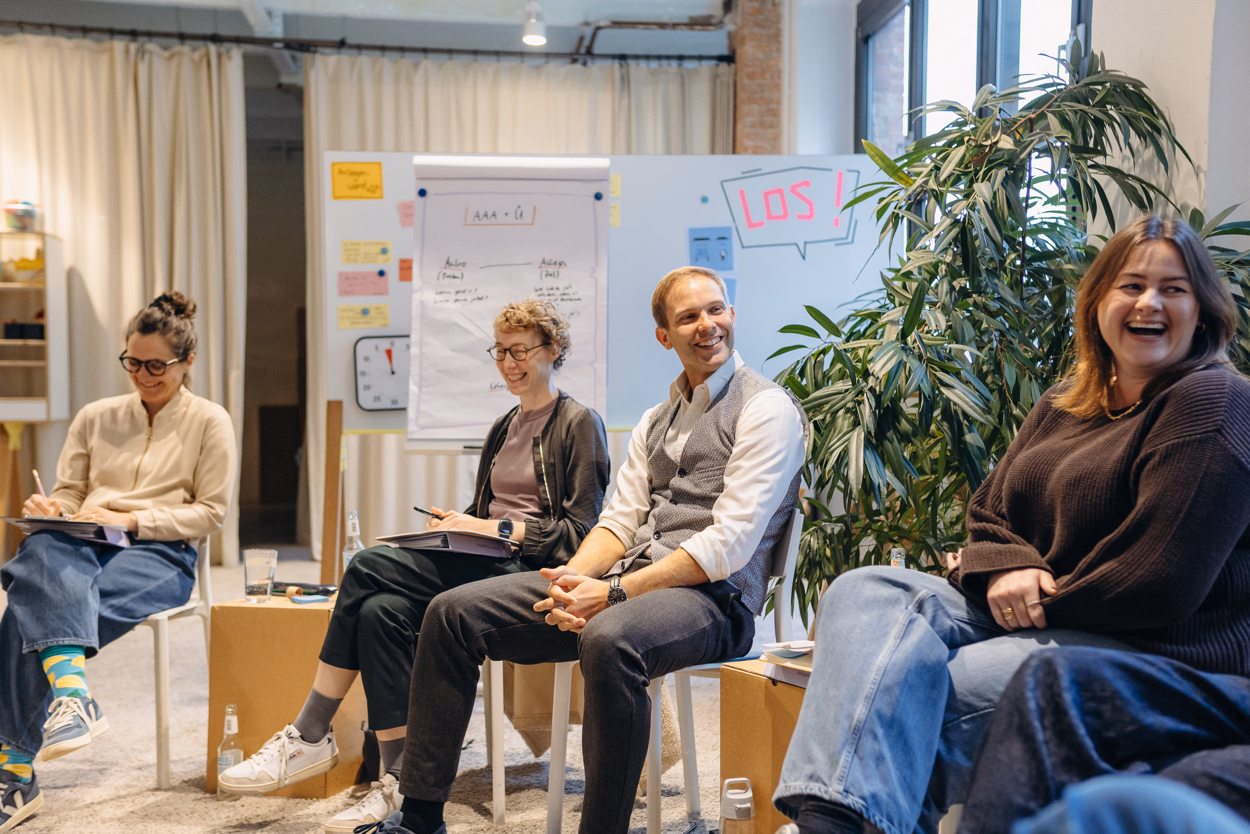 Kevin Rassner (mit Weste) lacht mit Teilnehmenden in einer Workshop-Runde, im Hintergrund Flipchart und Whiteboard mit „LOS!“