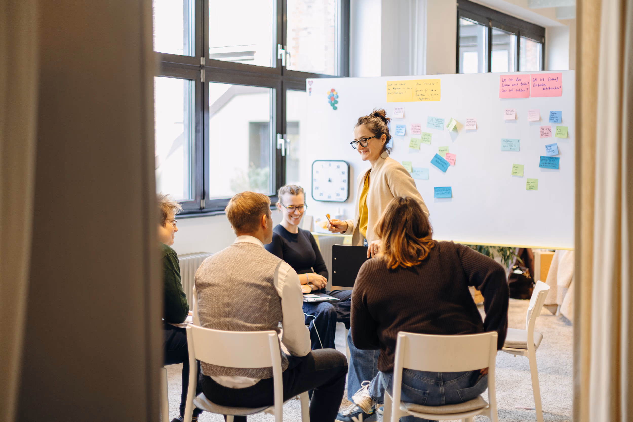 Workshopsituation: Moderatorin im Gespräch mit einer Gruppe im Stuhlkreis vor einem Whiteboard mit Moderationskarten